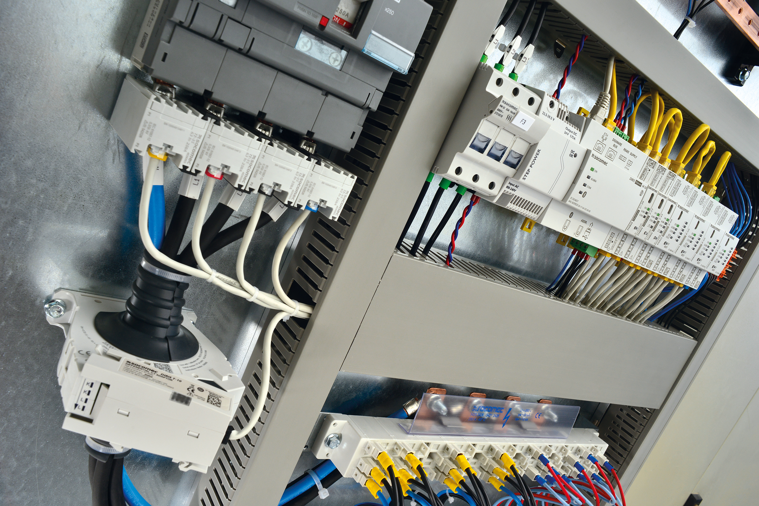 Nahaufnahme eines geöffneten Schaltschrankes mit SOCOMEC-Komponenten: Leistungsschalter/Schutzgeräte, Steuer- und Kommunikationsmodule sowie verdrahtete Klemmen- und Verteilblöcke mit farbcodierten Leitungen.